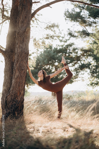 A woman practices a yoga balance pose outdoors, leaning against a tree in a calm, sunlit natural setting.