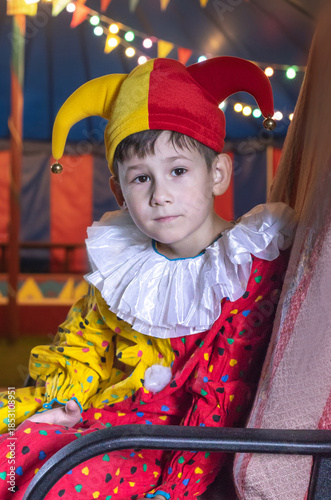 A boy in a bright jester costume and a two-tone hat with bells poses against the backdrop of a circus tent decorated with garlands