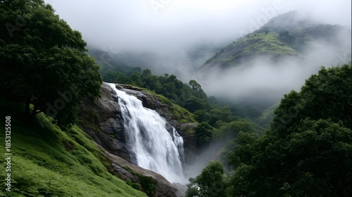 A majestic waterfall cascades down rocky slopes surrounded by lush green mountains veiled in ethereal mist