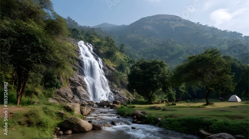 A majestic waterfall cascades down a lush green mountain into a rocky river with a tent set up in a nearby clearing