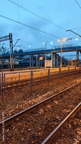 Railway tracks leading through an empty train station platform at dusk with overhead lines and pedestrian bridge. The image represents travel, transportation infrastructure, and urban transition