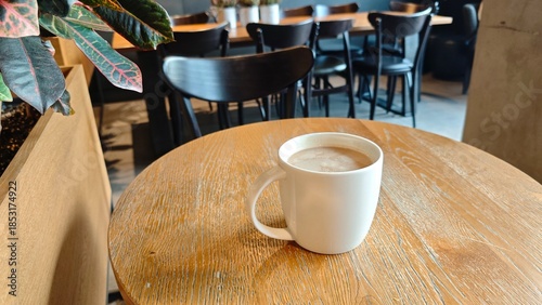 White coffee cup with hot drink on a wooden table inside a modern cafe with chairs and plants in the background. The image represents relaxation, cozy atmosphere, and everyday coffee break lifestyle
