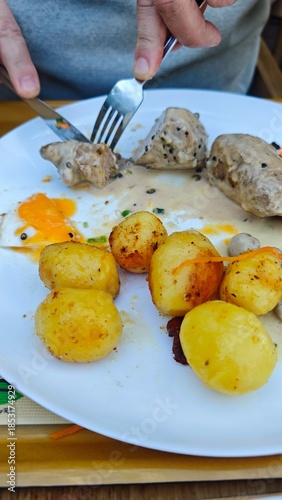 Plate with roasted potato and meat in creamy sauce being cut with fork and knife during a meal. The image represents dining, appetite, homemade food, and everyday eating experience