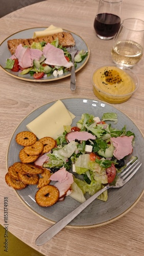 Plate with fresh salad, sliced ham, cheese, crackers, and fork served on a wooden table with wine and dip in the background. The image represents casual dining, shared meal, and relaxed home gathering