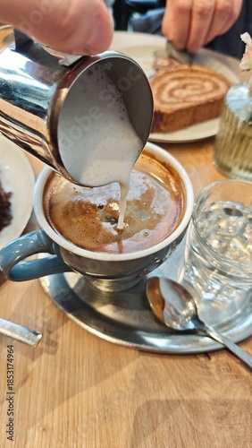 Cup of coffee with fresh milk foam being poured from a metal pitcher on a wooden cafe table. The image represents coffee ritual, craftsmanship, and cozy social moment