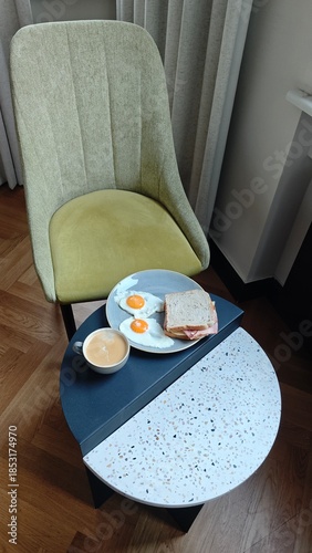 Plate with fried egg ham cheese sandwich and coffee on a small table in a cozy cafe setting. The image represents classic breakfast routine comfort food and relaxed morning lifestyle