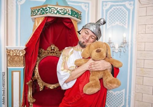 Funny smiling man weared king costume holds a teddy bear shows parody contrast