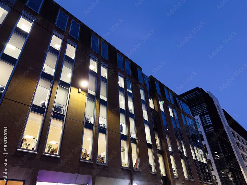Naklejka premium Modern office building in Manchester UK with illuminated windows at dusk and blue evening sky