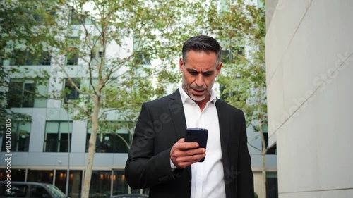 A focused businessman intently checking messages on his smartphone while walking outdoors in formal attire, showcasing a moment of modern communication and engagement in a professional setting.