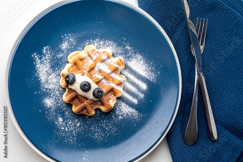 Belgian waffles or dessert with white cream and blueberries sprinkled with powdered sugar on a blue plate with napkin and cutlery. Top view. High quality photo