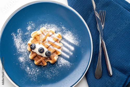 Belgian waffles or dessert with white cream and blueberries sprinkled with powdered sugar on a blue plate with napkin and cutlery. Top view. High quality photo