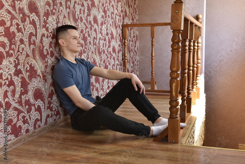 oung man sitting on wooden floor near staircase inside home, resting and thinking in calm domestic interior with natural light and casual atmosphere