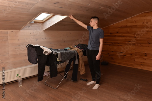 Young man opening roof window in attic room while drying clothes indoors, everyday home routine, casual lifestyle moment in wooden interior