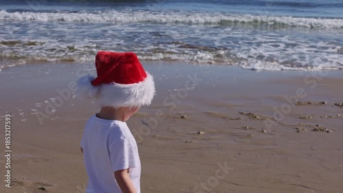 Little happy boy wearing red Santa hat running and jumping on a sandy beach near sea. Christmas holidays concept. 