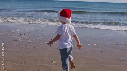 Little boy wearing red Santa hat running, jumping on a sandy beach and falling down near sea.