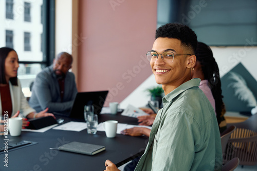 Confident young businessman smiling at camera while attending team meeting in boardroom at office