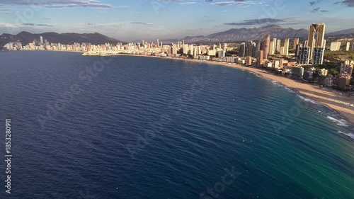 aerial calm over sweeping ocean curve at dawn with glassy water, subtle ripples, distant city silhouette