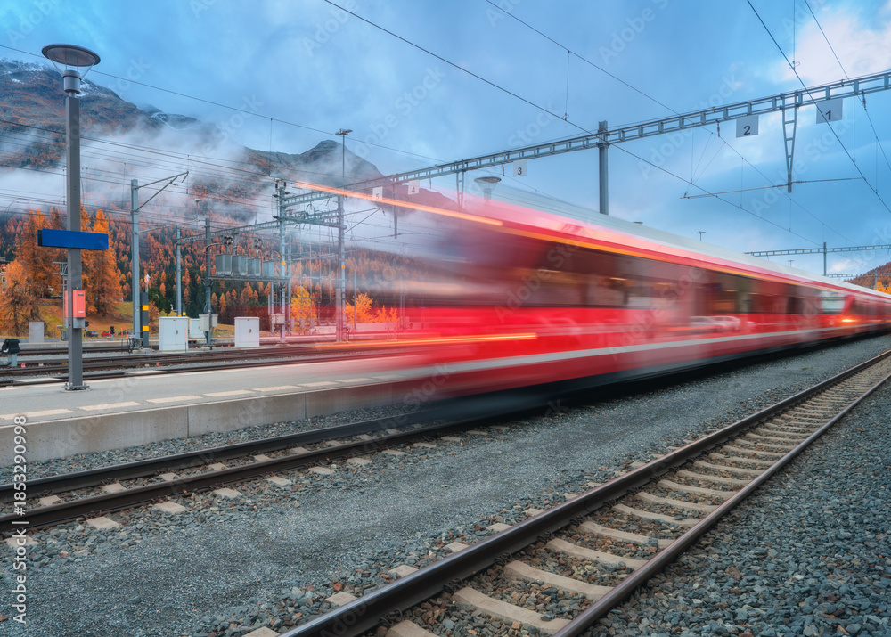 Naklejka premium Blurred red passenger train passing mountain railway station in Swiss Alps at dusk. Moving high-speed train. St. Moritz, Switzerland. Bernina Express. Railway platform and cloudy sky. Rail transport
