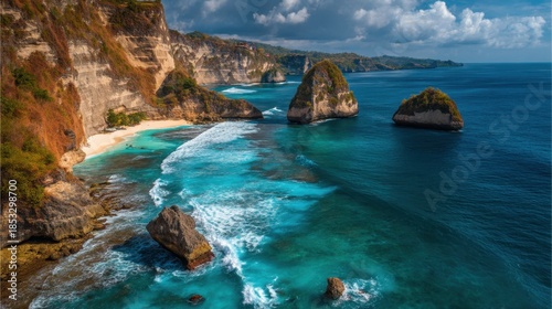 Cliffs rise steeply from a beach with soft sand and clear water. Rocky formations dot the coastline under a bright blue sky. The scene shows waves breaking along the shore.