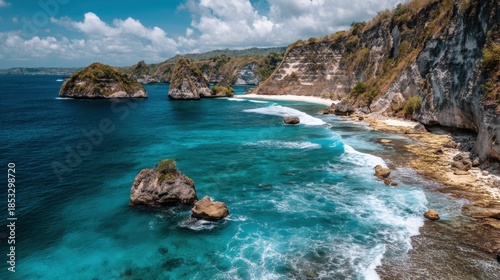 Waves crash on the rocks along a tropical coastline. Green hills rise in the background while fluffy clouds float above a sunny sky. The water is clear and blue.
