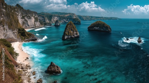 Cliffs rise steeply from a beach with soft sand and clear water. Rocky formations dot the coastline under a bright blue sky. The scene shows waves breaking along the shore.