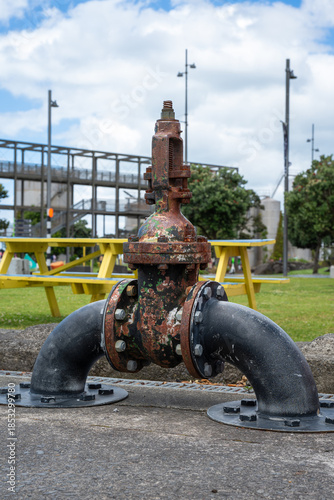 A vintage iron water valve with peeling red paint sits on a rusted pipeline, showing years of industrial wear and tear.