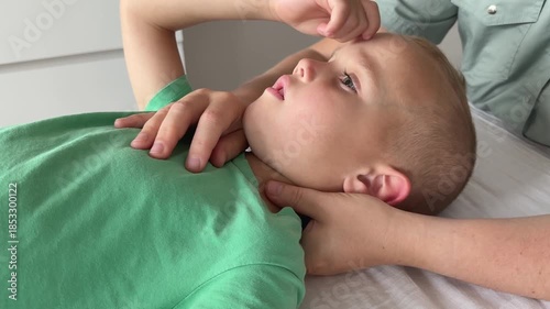 Side view of young child lying on white therapy couch during osteopathy session. Doctor holds one hand on neck and another on chest, while boy scratches forehead, blinks, and looks up at therapist.