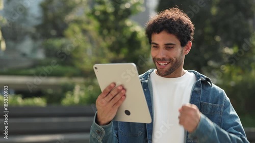 Portrait of emotional Hispanic man with digital tablet expresses joy by winner gesture with fist in park. Latin guy wins online lottery by mobile device in summer megapolis block