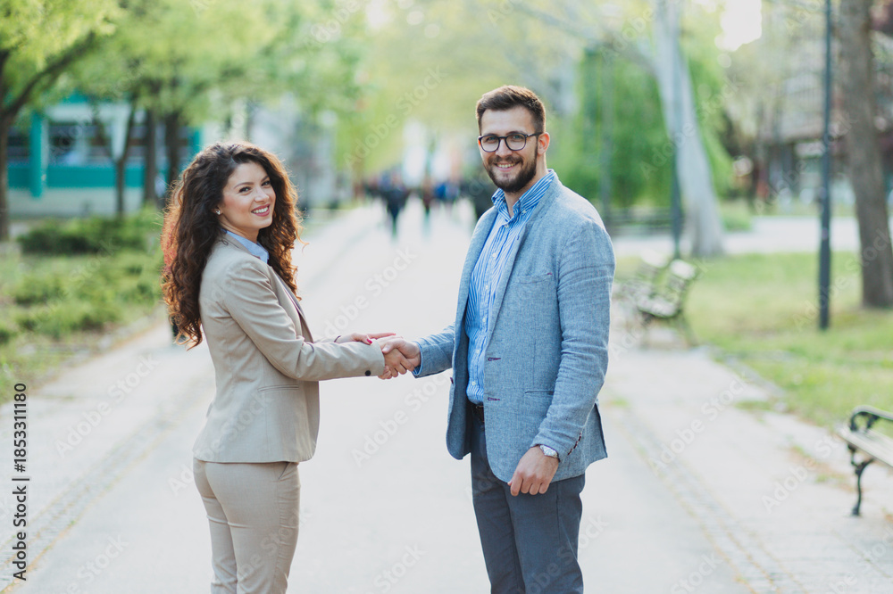 Obraz premium Professional man and woman in suits shaking hands in a public park, representing a business deal, academic partnership, or formal agreement outdoors.