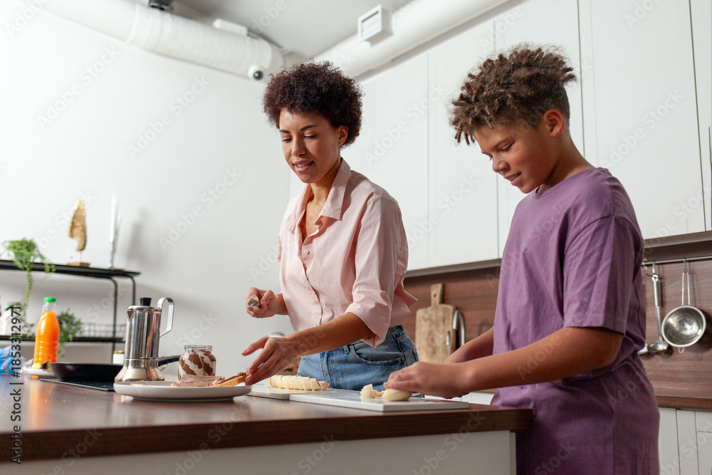Fototapeta premium African-American woman and her son prepare chocolate butter and banana sandwiches in the kitchen, while teenager helps his mother prepare breakfast at home.