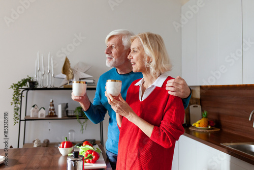 elderly couple of seniors drink coffee in the kitchen and smile, old woman and man hold cups of tea and hug at home
