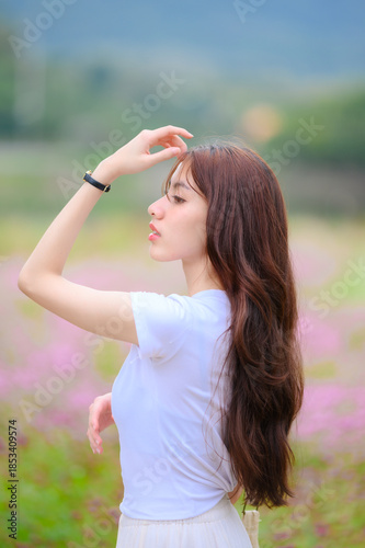 Beautiful young Asian woman with long wavy brown hair posing in a blooming pink flower field. Soft focus background, side profile, romantic lifestyle and beauty portrait.
