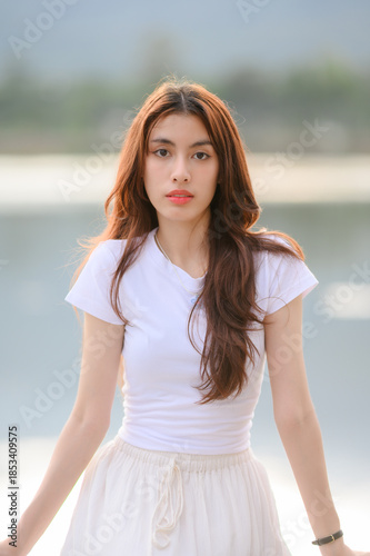 Beautiful young Asian woman with long wavy brown hair looking at camera, wearing white t-shirt against blurred lake background. Natural soft light beauty portrait.