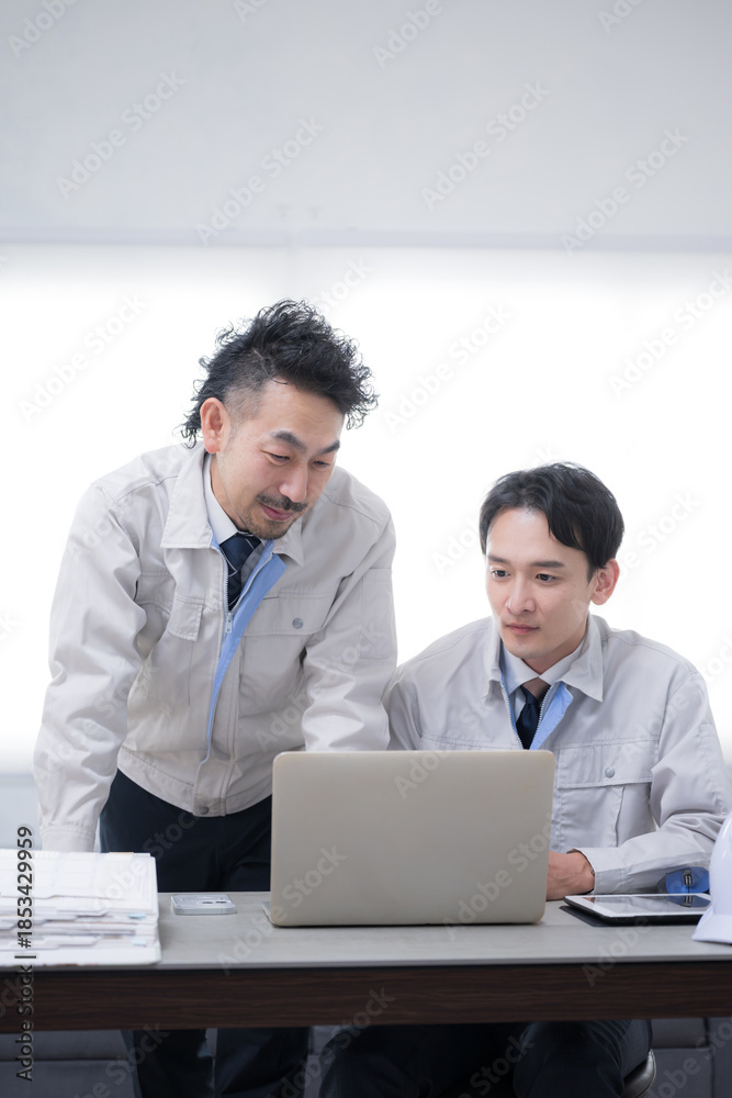 Fototapeta premium Two businessmen in work clothes talking while looking at a computer Image of DX and IT adoption in manufacturing and other industries