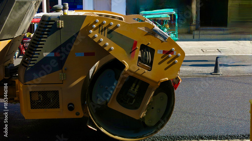 Road roller compresses fresh asphalt during city street repairs. Heavy construction machine works on urban road maintenance, showing detailed industrial equipment and active infrastructure repair.