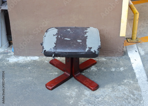 A footstool on concrete floor. The stool's padded top is in poor condition, showing significant wear where the brown faux leather covering has peeled away, exposing the grey material beneath.