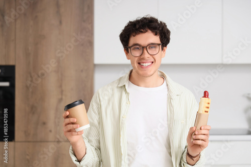Young man with tasty hot dog and coffee cup in kitchen