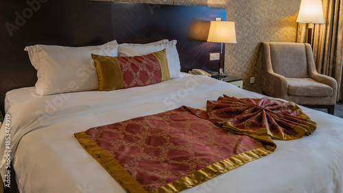 The double bed is beautifully made. A red and gold bedspread is decoratively laid on white sheets. Pillows at the headboard. A chair in the corner. The floor lamp is lit. Bedroom. Hotel. 