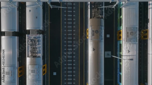 Aerial view of commuter trains parked at a station in Auckland, New Zealand. The trains are silver and blue, and the tracks are black. The scene shows the tops of the trains and the tracks below.