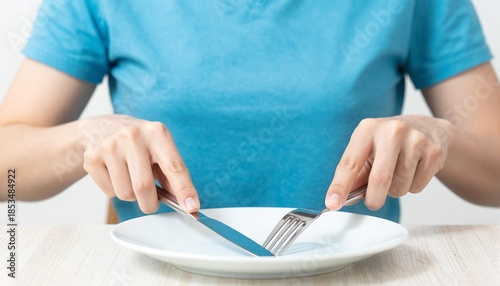 Person in blue shirt holding knife and fork poised over an empty white plate on a light wooden table, suggesting waiting, hunger, dieting or mealtime