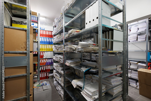An interior view of a document archive room with metal shelving units filled with stacks of paper files, folders, and colorful ring binders