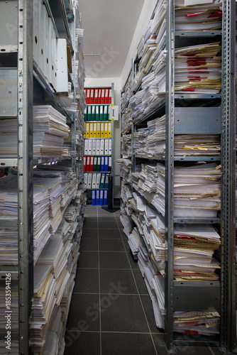 An interior view of a document archive room with metal shelving units filled with stacks of paper files, folders, and colorful ring binders