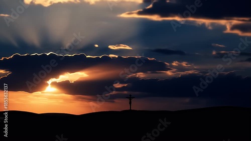 Dramatic sunset with silhouette of person on a cross against layered dark moody clouds illuminated by golden warm light and rays of sunshine