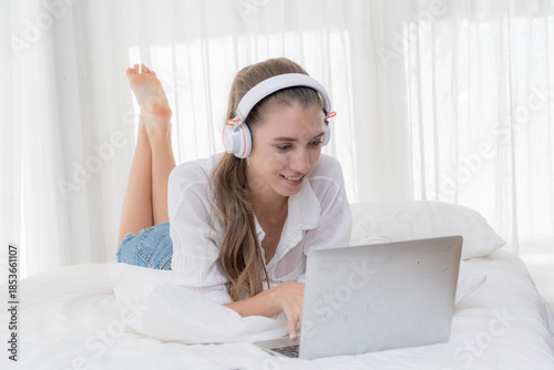 Happy caucasian woman blonde long hair enjoy her laptop with headphone on a white bed, representing cozy home entertainment, online study, or comfortable remote working.