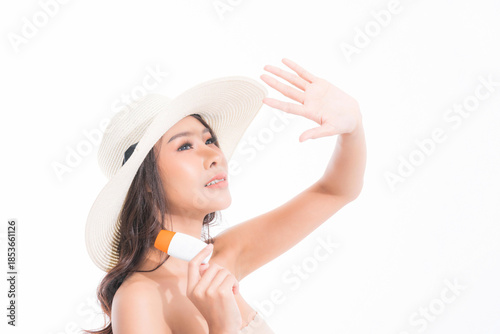 Young beautiful asian woman in a white sun hat shielding her eye from the sun with her hand, holding sunscreen. Skincare and summer concept at studio shot isolated on white background.