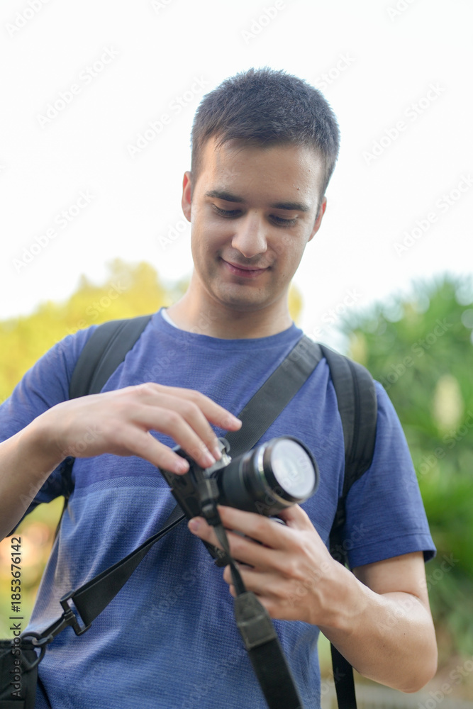 Fototapeta premium A man is holding a camera and smiling. He is wearing a blue shirt and a backpack. The camera is attached to a strap around his neck