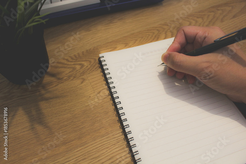 Close up of hand writing on blank spiral notebook with pen on wooden desk. Lifestyle of businessman or student taking notes on paper at home office.
