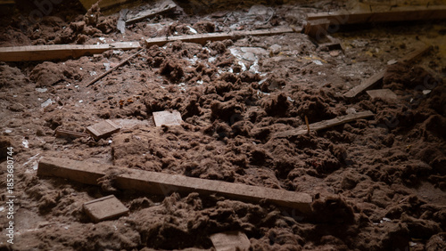 Dust covers the floor where wooden beams lie scattered among debris in a neglected building with light filtering through gaps, revealing a disordered space.