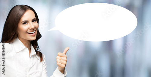 Portrait image - expressive happy smile business woman in white shirt show point thumb up hand sign gesture. Businesswoman stand on blurred office background with empty free space mockup speech bubble