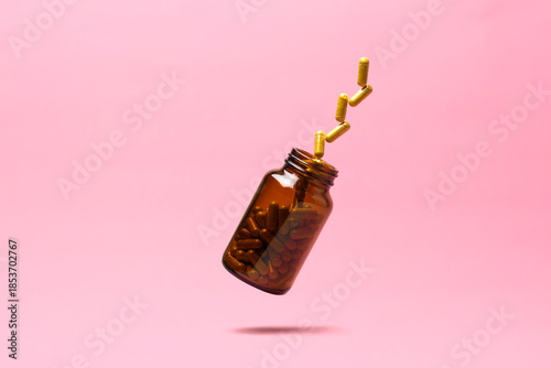 Amber supplement bottle in midair releases golden capsules on pink background, clean copy space for modern wellness branding.